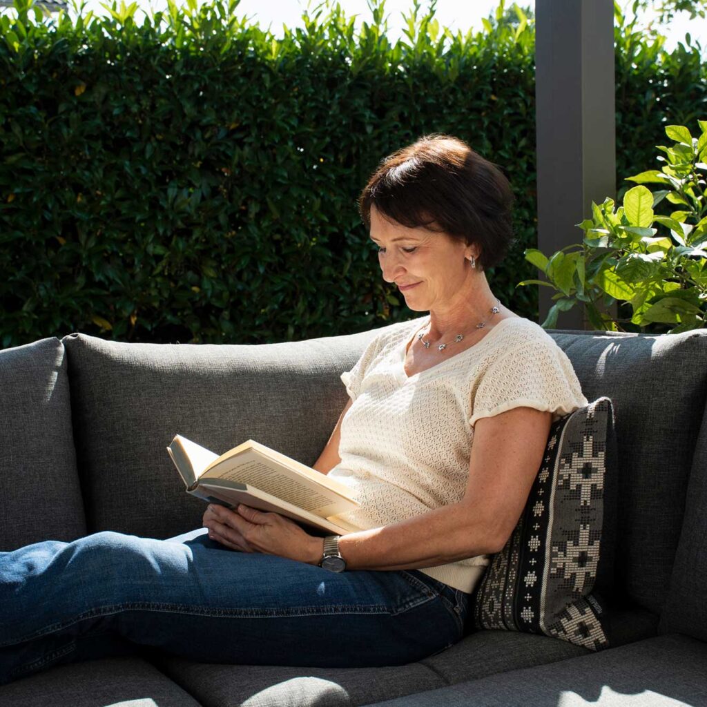 frau sitzt auf grauem outdoor-sofa vor hoher heckenpflanze. sie liest ein aufgeschlagenes buch und hält es mit beiden händen.