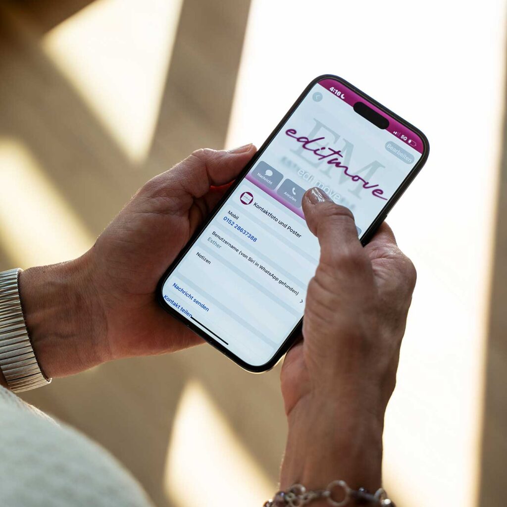 zwei hände halten smartphone mit geöffneter editmove-app-kontaktseite, schriftzug editmove, mobil 0152 26837388, benutzername esther, sonnenlichtflecken im hintergrund.