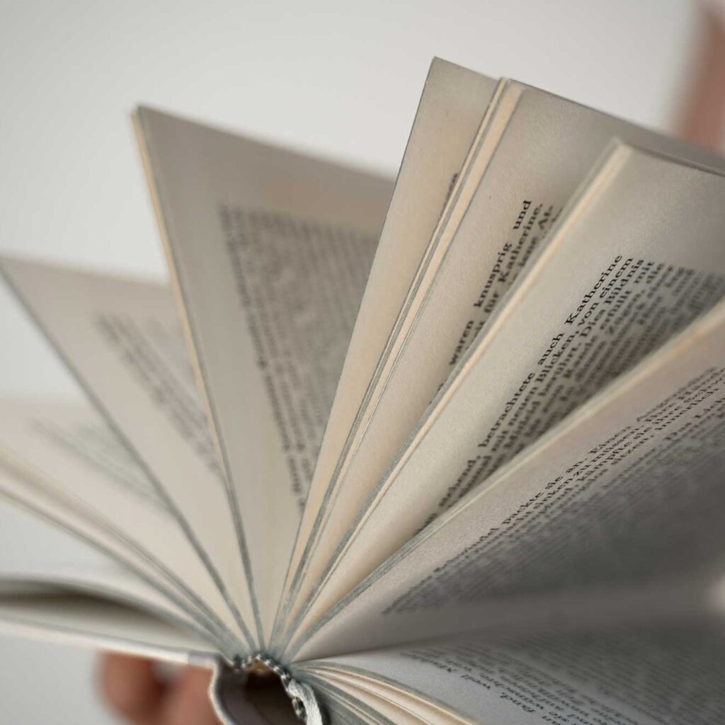 person hält aufgeklapptes buch vor weißem hintergrund, seiten fächerartig gespreizt und textzeilen sichtbar.