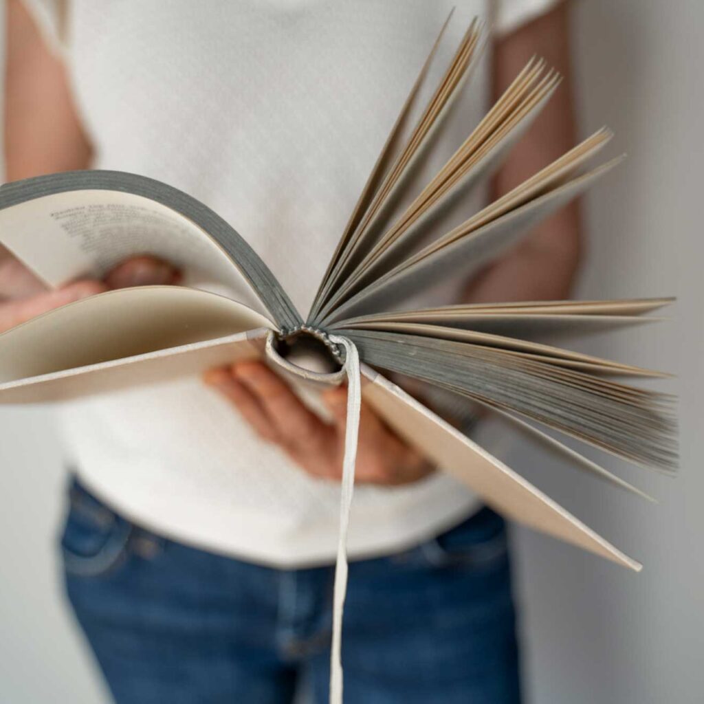 person hält aufgeschlagenes buch mit fächerartig gespreizten seiten vor weißem hintergrund.